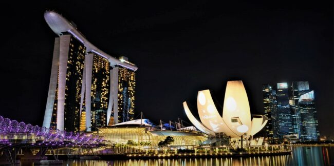 Singapur-Next Step Asia Nachtaufnahme einer modernen Stadt mit markanten, beleuchteten Gebäuden. Im Vordergrund spiegelt sich das Licht auf dem Wasser. Links steht ein großes Gebäude mit drei Türmen, die von einer länglichen Struktur auf dem Dach verbunden sind. Daneben ist ein futuristisches, blütenförmiges Gebäude mit hellen Projektionen zu sehen. Im Hintergrund stehen weitere Wolkenkratzer mit hell erleuchteten Fenstern