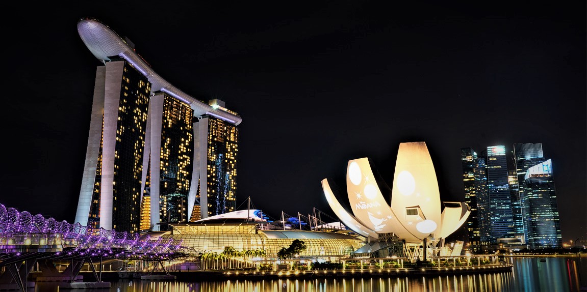 Singapur-Next Step Asia Nachtaufnahme einer modernen Stadt mit markanten, beleuchteten Gebäuden. Im Vordergrund spiegelt sich das Licht auf dem Wasser. Links steht ein großes Gebäude mit drei Türmen, die von einer länglichen Struktur auf dem Dach verbunden sind. Daneben ist ein futuristisches, blütenförmiges Gebäude mit hellen Projektionen zu sehen. Im Hintergrund stehen weitere Wolkenkratzer mit hell erleuchteten Fenstern