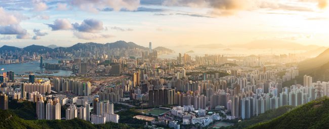 Kowloon View point sunset Panoramablick auf eine dicht bebaute Stadt mit zahlreichen Hochhäusern, die sich bis zum Wasser und den Bergen im Hintergrund erstreckt. Die Sonne scheint durch leichte Wolken