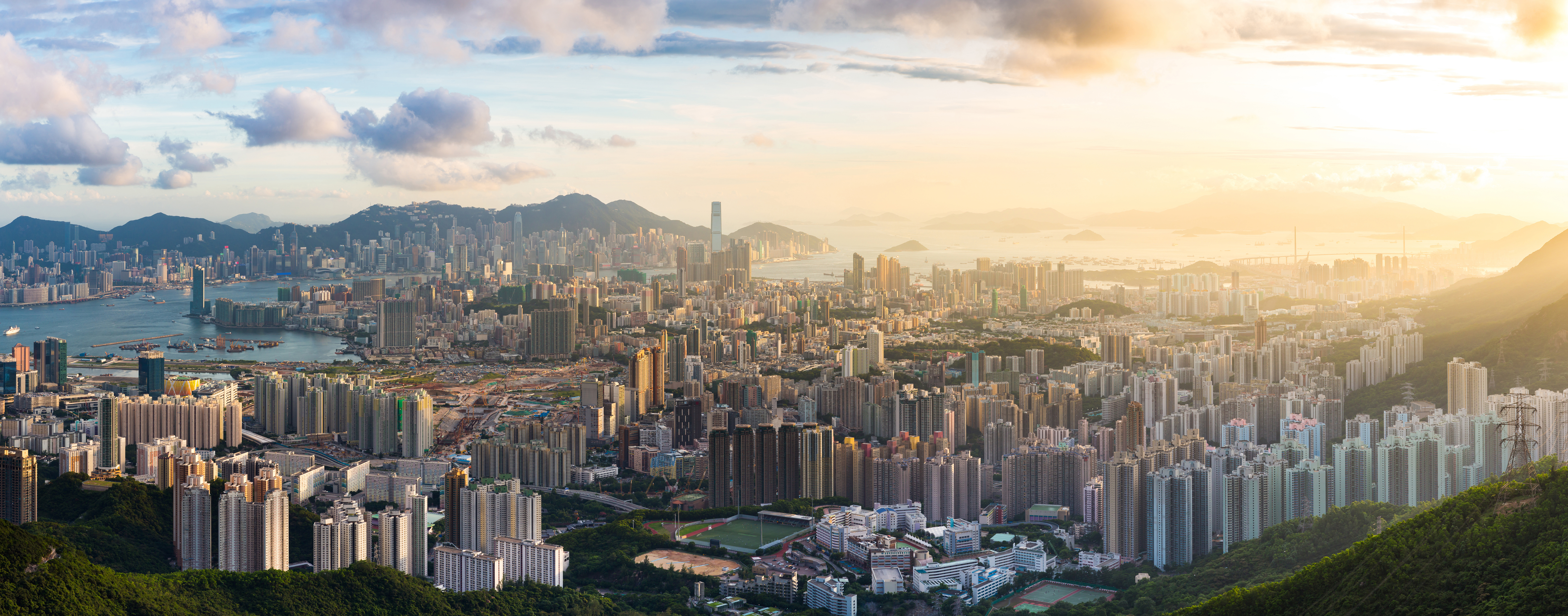 Kowloon View point sunset Panoramablick auf eine dicht bebaute Stadt mit zahlreichen Hochhäusern, die sich bis zum Wasser und den Bergen im Hintergrund erstreckt. Die Sonne scheint durch leichte Wolken