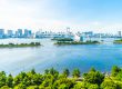 Panoramablick auf die Rainbow Bridge in Tokio bei klarem Himmel, umgeben von Wolkenkratzern, Wasserflächen und grüner Vegetation im Vordergrund