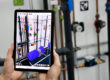 A person holds a tablet in front of an industrial pipe installation. The screen shows an augmented reality display that overlays the real environment with colored virtual components such as pumps and valves