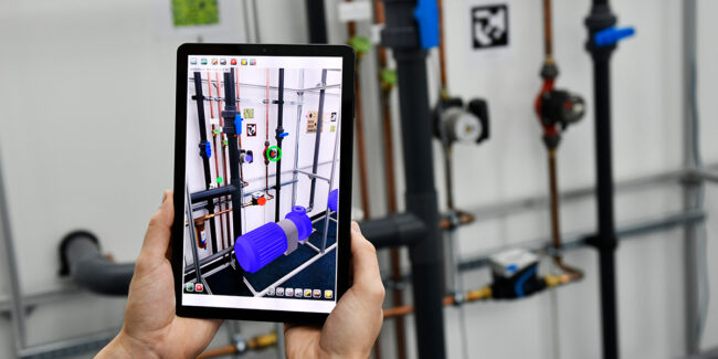 Framence_Blog_AR A person holds a tablet in front of an industrial pipe installation. The screen shows an augmented reality display that overlays the real environment with colored virtual components such as pumps and valves