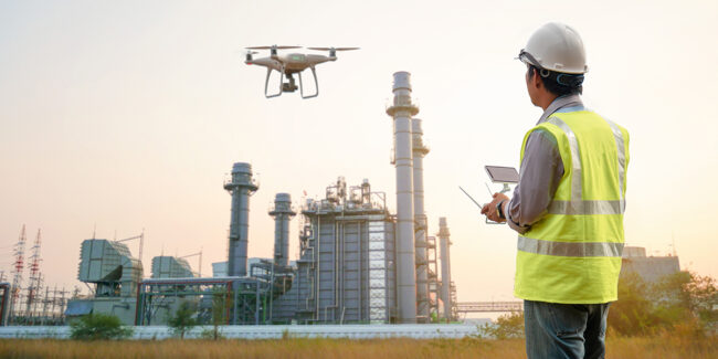 Framence_Blog_Drohne An engineer in a helmet and yellow safety vest uses a remote control to operate a drone in front of a large industrial plant at sunset