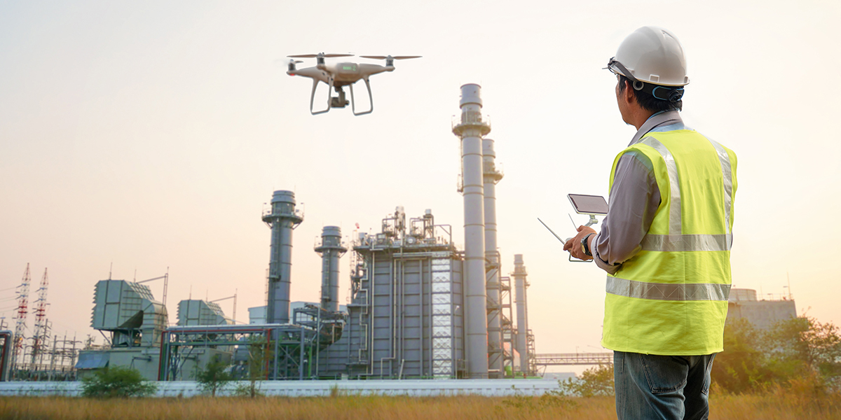 Framence_Blog_Drohne An engineer in a helmet and yellow safety vest uses a remote control to operate a drone in front of a large industrial plant at sunset