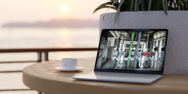 Framence_Blog_Neue_Arbeitswelt Laptop stands on a round wooden table next to a white coffee cup. The screen shows a complex installation of pipes, valves and measuring devices in a technical room. In the background, a blurred landscape with water and sunset, some green plants.